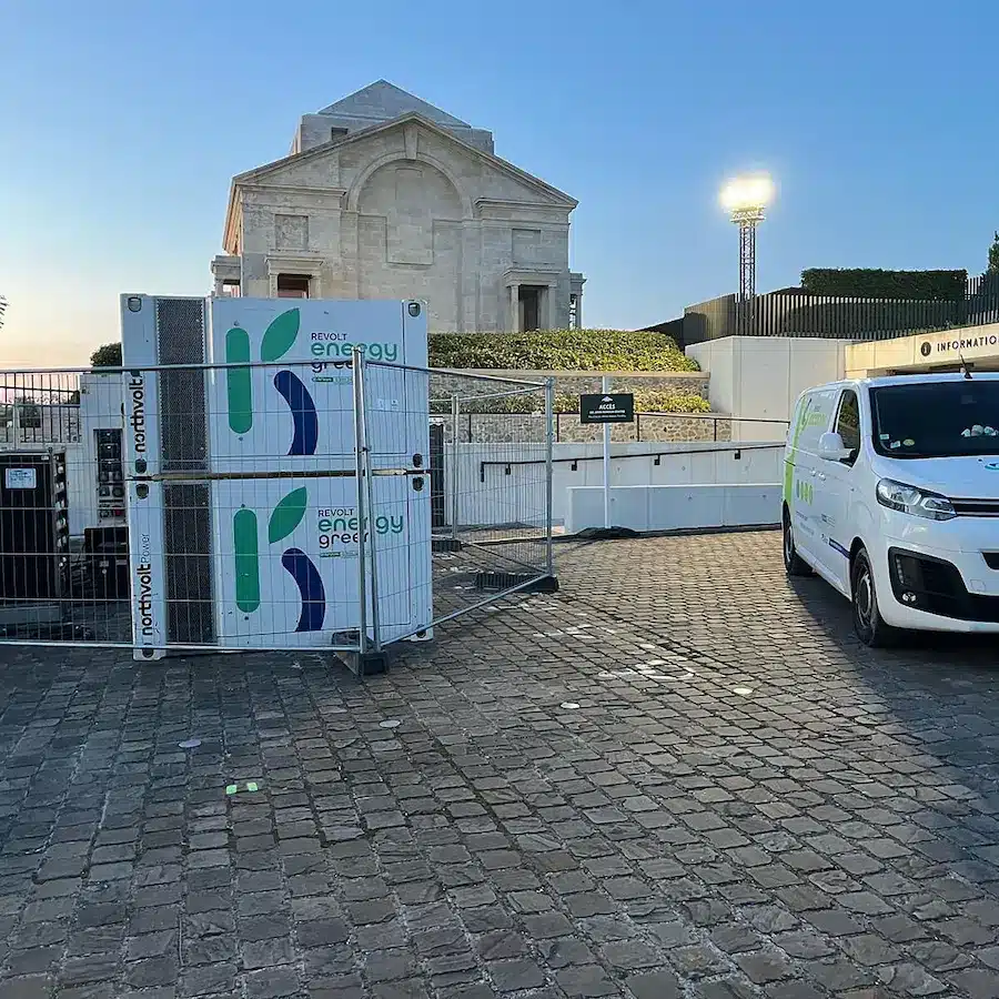 Unités de stockage d'énergie par batteries Revolt Energy Green (technologie Northvolt) installées sur un parking pavé devant un monument historique, avec un fourgon d'intervention Revolt.