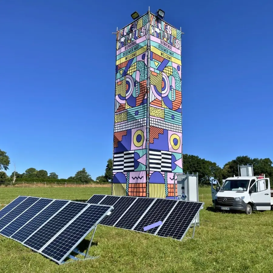 Alignement de panneaux solaires photovoltaïques au sol devant un totem décoré aux couleurs du festival des Vieilles Charrues, avec une armoire de distribution électrique et un fourgon technique.