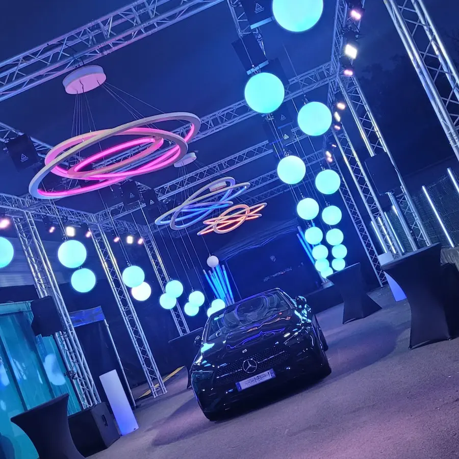 Éclairage événementiel design avec sphères lumineuses bleues et lustres circulaires néon surplombant une voiture de luxe Mercedes lors d'une soirée de lancement.